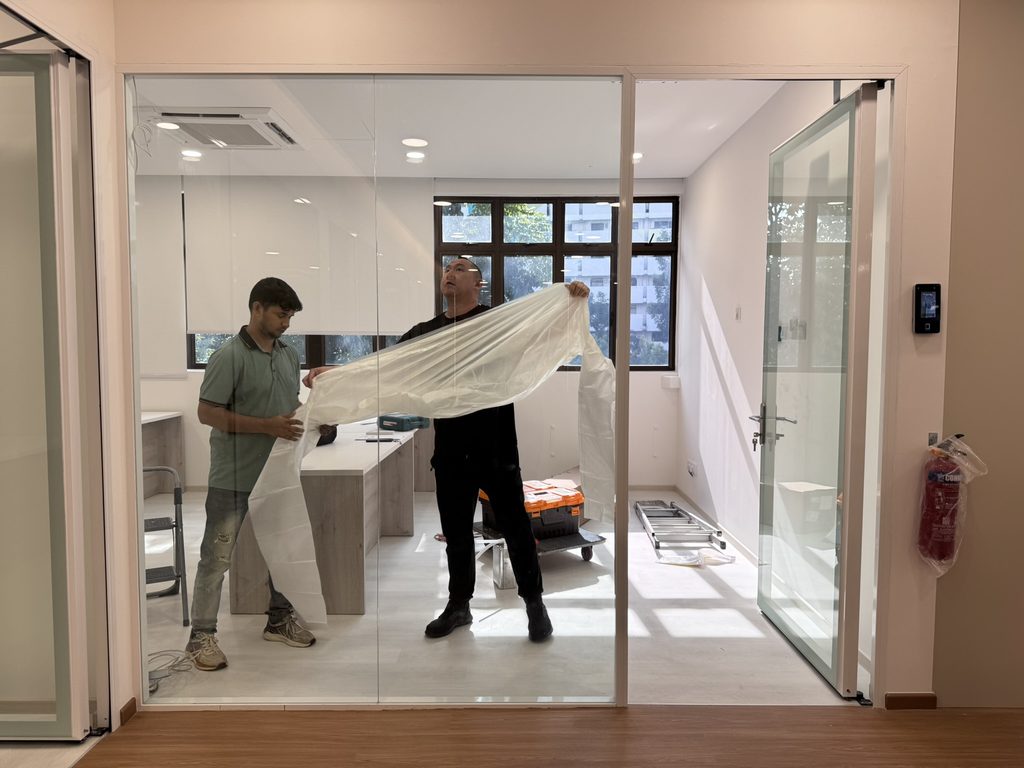 Installers handling a sheet of smart film inside a glass-partitioned room before applying it to the existing glass